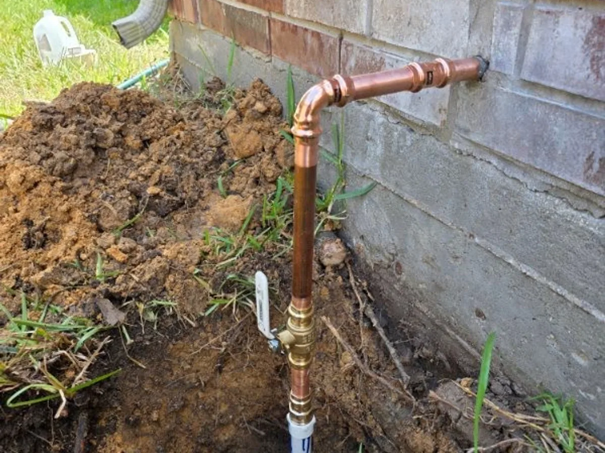 New copper water line riser entering a brick wall above the foundation, with a brass ball valve shutoff and a transition to buried poly pipe.